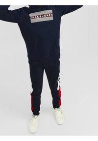 Sudadera azul marino con un bolsillo frontal y un logo a rayas en blanco, rojo y azul. Combinada con pantalones jogger a juego que tienen acentos laterales en rojo y blanco.