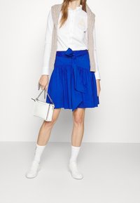 Blå skaterkjol med midjeband, vit skjorta med knappar, ljusbeige stickad väst, vita sneakers och en liten vit handväska med kedjerygg.