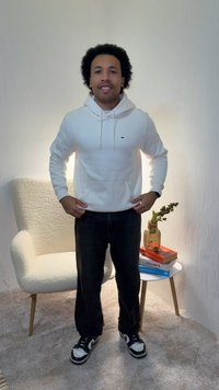 Biela mikina s kapucňou a predným vreckom, skombinovaná s čiernymi džínsami a čierno-bielymi teniskami. Neutrálne pozadie s textúrovanou stenou.