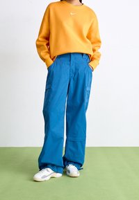 Orange sweatshirt med rund hals och vit logotyp, kombinerat med blå cargobyxor med fickor. Vita sneakers fulländar outfiten.