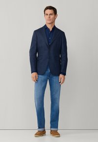 Mann steht und trägt ein dunkelblau gemustertes Sakko, ein navyblaues Hemd, hellblaue Jeans mit Umschlägen und braune Wildlederschuhe vor einem einfarbigen grauen Hintergrund.