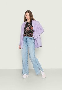 Blazer oversized lilás, top preto com padrão floral, jeans de perna larga azul claro e ténis brancos com detalhes. Modelo está contra um fundo neutro.