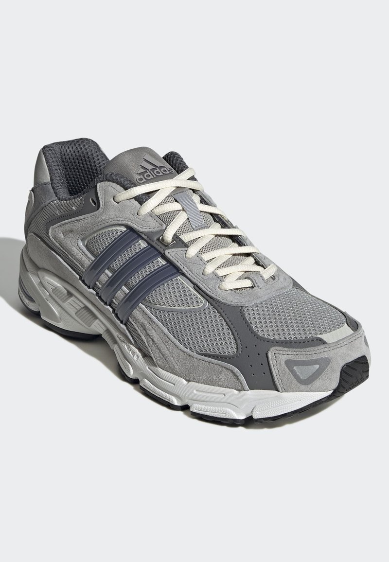 Zapatilla deportiva gris con material de malla y ante, que presenta tres rayas gris oscuro, cordones blancos y una suela de goma texturizada.