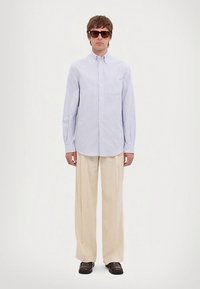 Vīrietis, kurš valkā pārāk lielu gaiši zilu svītrainu krekls ar pogām, platus bežīgas bikses, tumši brūnus apavus un lielas tumšas saulesbrilles, stāv uz baltā fona.