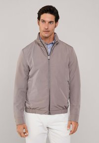 Giacca leggera color taupe con zip e colletto alto, orlo elasticizzato. Tessuto liscio e lucido con tasche laterali e design minimalista.