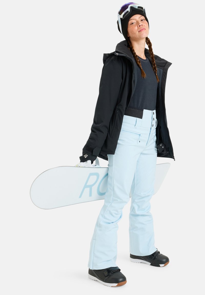 Snowboardzista w czarnej kurtce, czapce beanie i ciemnej koszulce, z jasnoniebieskimi spodniami narciarskimi i czarnymi butami, trzymający białą deskę snowboardową.