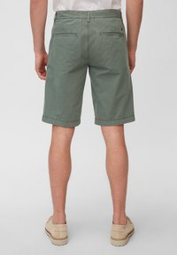 Miestne zelené bavlnené šortky s pohodlným strihom, s jednoduchou textúrou, dvoma zadnými vreckami a zaobleným lemom. Nosené s svetlými topánkami.