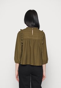 Olivgrüne, strukturierte Bluse mit Rüschenschultern, Dreiviertelärmeln und einem Schlüsselloch-Detail an der Rückseite. Lockere Passform mit besticktem Design.