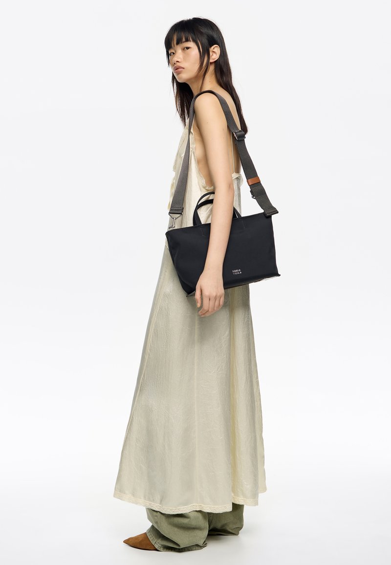 Czarna prostokątna torba z dwoma uchwytami i regulowanym paskiem. Gładka tekstura, minimalistyczny design i subtelne oznakowanie z przodu.