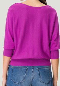 Maglione viola lavorato a maglia con scollatura ampia, maniche a tre quarti e polsini a costine, abbinato a jeans in denim blu.