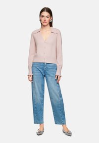 Helle rosa Strickjacke mit V-Ausschnitt, Knopfverschluss, langen Ärmeln und Volantmanschetten, kombiniert mit hellblauen geraden Jeans und grauen Ballerinas.