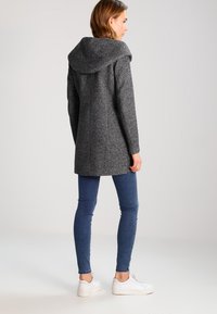 Manteau en mélange de laine gris avec une grande capuche drapée, une coupe cintrée et des coutures verticales, associé à un jean skinny bleu et des baskets blanches.