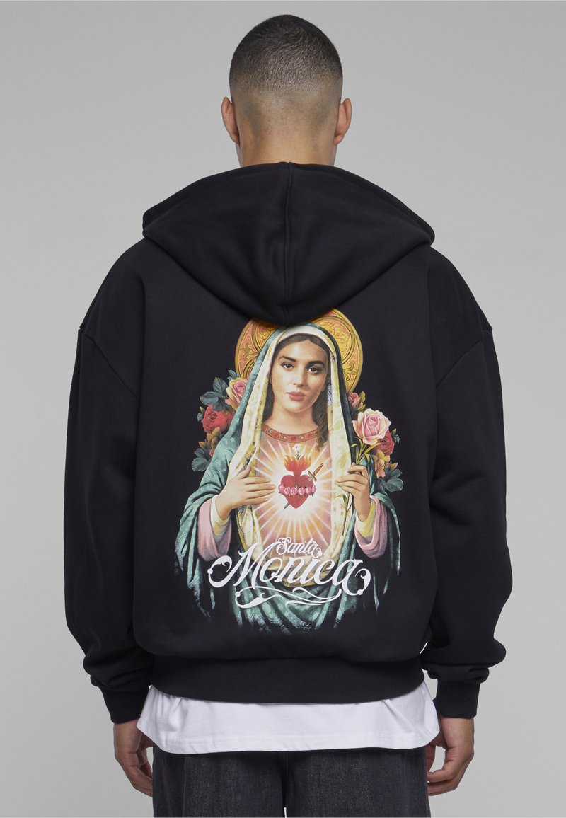 Mies, jolla on päällään musta huppari, jonka takana on värikäs Pyhän Sydämen ja Pyhän Monican kuva, pitäen kädessään vaaleanpunaista ruusua.