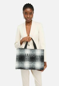 Tote bag i sort/hvid plaid med tekstureret stof, med sorte håndtag og "LIU JO" logo på forsiden. Rektangulær form.