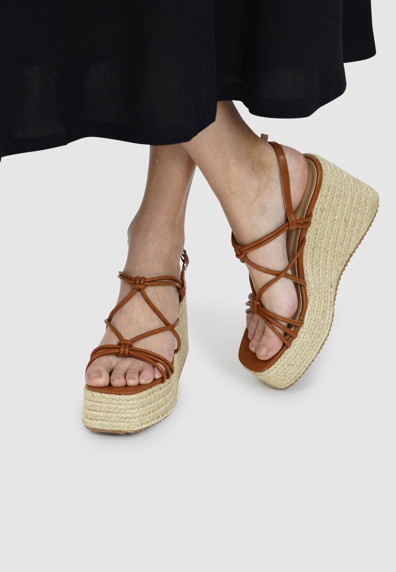 Bruine leren wedge sandalen met kruislings straps, een geweven touwzool en een plateauontwerp. Het teengebied is open en toont blote voeten.
