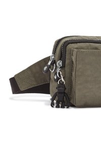 Khaki fabric bag with two zippered compartments, black accents, and a small black monkey charm attached. Adjustable strap on one side.