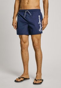 Tengeri úszónadrág fehér "Pepe Jeans" logóval, elasztikus derékpánttal és zsinórral, könnyű anyagból; fekete strandpapuccsal viselve.