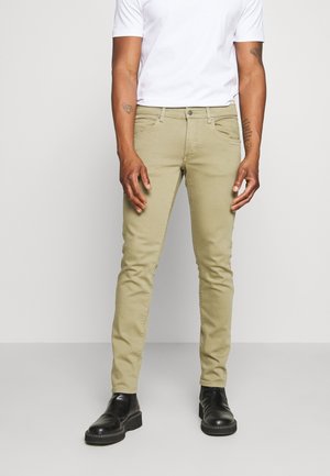 Chinos en coton beige avec une coupe slim, dotés d'une fermeture éclair, d'un design à cinq poches et de coutures subtiles. Portés avec une chemise blanche et des bottes noires.