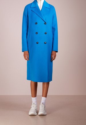 Person wearing a knee-length blue double-breasted coat with black buttons, white sneakers, and white socks, standing against a plain background.