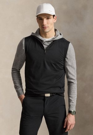 Mand iført hvid baseballkasket, grå hættetrøje under sort ærmeløs lynlåsvest, sorte bukser og ur på venstre håndled mod en neutral baggrund.