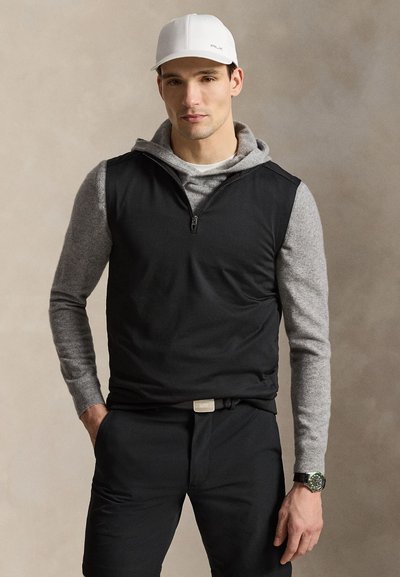 Homme portant une casquette de baseball blanche, un sweat à capuche gris sous une veste sans manches noire zippée, un pantalon noir et une montre au poignet gauche sur un fond neutre.