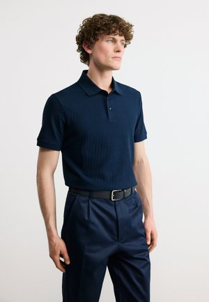 Jeune homme aux cheveux bouclés portant un polo bleu marine texturé rentré dans un pantalon bleu marine avec une ceinture noire, regardant sur le côté.