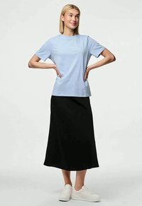 Mulher de pé com as mãos nos quadris, vestindo uma t-shirt azul clara, saia midi preta e ténis brancos, sorrindo levemente, em fundo simples.