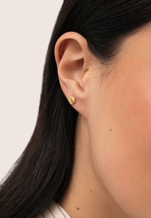 Primer plano de una oreja con cabello oscuro que lleva un pequeño pendiente de botón en forma de rosa de oro contra un fondo liso.