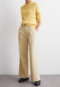 Persona che indossa un maglione a maglia gialla e pantaloni a vita alta beige a gamba larga con mocassini neri, in piedi contro un muro bianco.