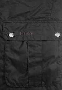Zwarte jas met een voorzak, Barbour-logo in bruin geborduurd en zilveren drukknoopjes. Glad, matte textuur. Eenvoudig ontwerp.