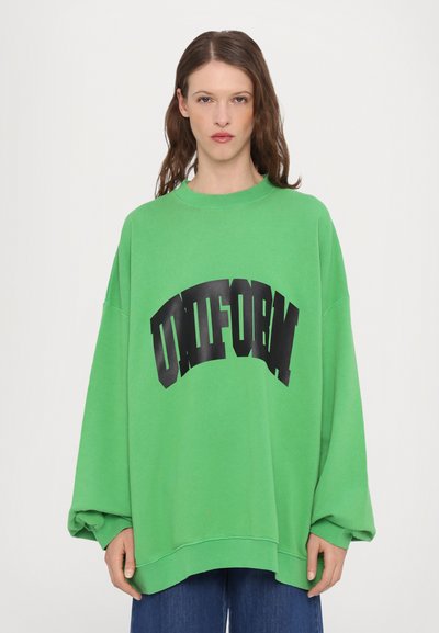 Zeleni oversized dukserica od pamuka, sa velikim, crnim zakrivljenim natpisom "UNIFORM" na prednjoj strani. Dugi rukavi i opušteni kroj.