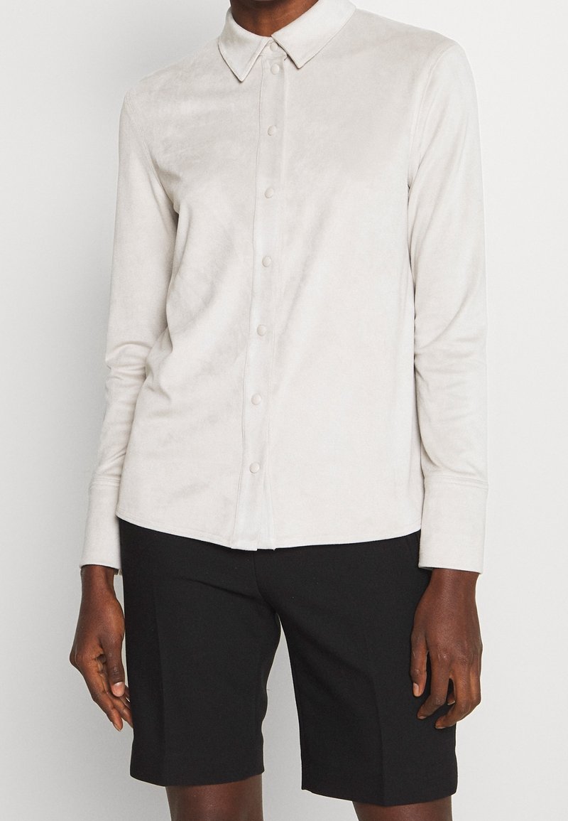 Personne portant une chemise beige clair à manches longues boutonnée associée à un short noir longueur genou sur un fond uni.