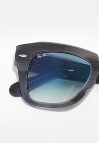 Ochelari de soare Ray-Ban cu un cadru mat de culoare albastru închis, lentile gradient de culoare albastră și un accent metalic argintiu aproape de braț. Design pătrat.