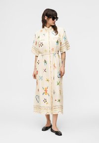 Γυναίκα που φοράει μπεζ midi φόρεμα με πολύχρωμα αφηρημένα σχέδια, με ζώνη στη μέση, κοντά μανίκια, μαύρες μπαλαρίνες και μαύρα γυαλιά ηλίου, στέκεται μπροστά σε λευκό φόντο.