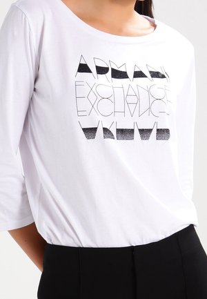 Biała koszula z długim rękawem z czarnym geometrycznym napisem "Armani Exchange", noszona z czarnymi spodniami.