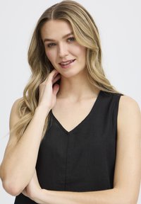 Blonďatá žena s vlnitými vlasy se jemně usmívá, má na sobě bezramínkový černý top s V výstřihem, ruka jí dotýká krku na bílém pozadí.