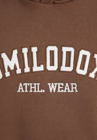 Sweatshirt marron avec le texte brodé blanc "HAMILODON ATHL. WEAR." Le texte présente une police en gras et stylisée avec une texture légèrement en relief.