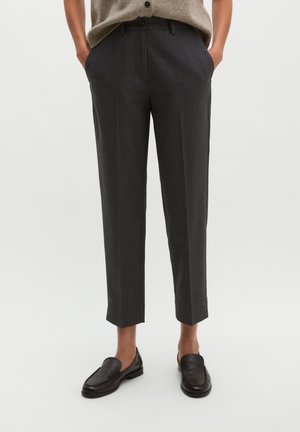 Person trägt dunkelgraue, schmal zulaufende Nadelstreifenhose und schwarze Loafers, Hände in den Taschen, steht vor einem schlichten, hellen Hintergrund.