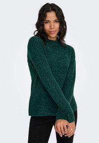 Zielony sweter z prążkowanego dzianiny z długimi rękawami, okrągłym dekoltem i luźnym krojem. Sweter ma subtelną fakturę i jest zestawiony z czarnymi spodniami.