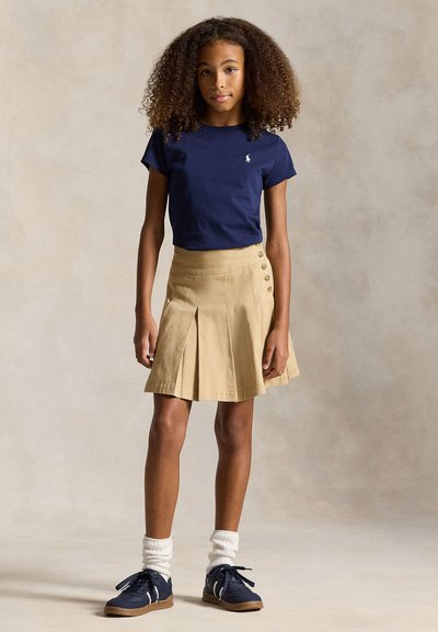 Mergina su garbanotais plaukais, vilkinti tamsiai mėlyną marškinėlį, smėlio spalvos plisuotą sijoną su sagomis, baltas kojines ir tamsiai mėlynas sportbačius, stovinti prieš paprastą foną.