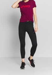 Camiseta burdeos de Asics con un logo en el frente, combinada con leggings negros y zapatillas deportivas negras con acentos rojos. Mangas cortas y diseño ajustado.