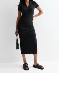 Schwarzes geripptes Midikleid mit Polokragen, kurzen Ärmeln, seitlichem Schlitz, kombiniert mit schwarzen Rutschsandalen und einer kleinen schwarzen Handtasche.