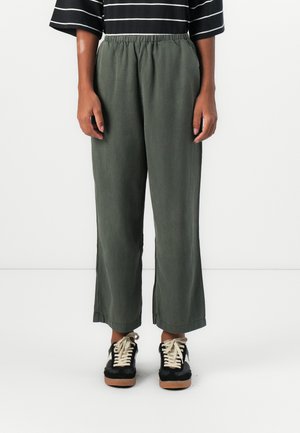 Green, wide-leg pants made of light fabric with an elastic waistband. Paired with black sneakers featuring beige laces and a textured sole.