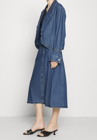 Jeansjacka med strukturerade axlar, draperade ärmar och kort design. Matchas med en lång jeanskänning med knapppdetaljer. Svarta sandaler med öppen tå.