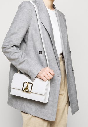 Personne portant un blazer gris clair et un pantalon beige, tenant un sac à main blanc avec un fermoir doré en forme d'abeille par la bandoulière.