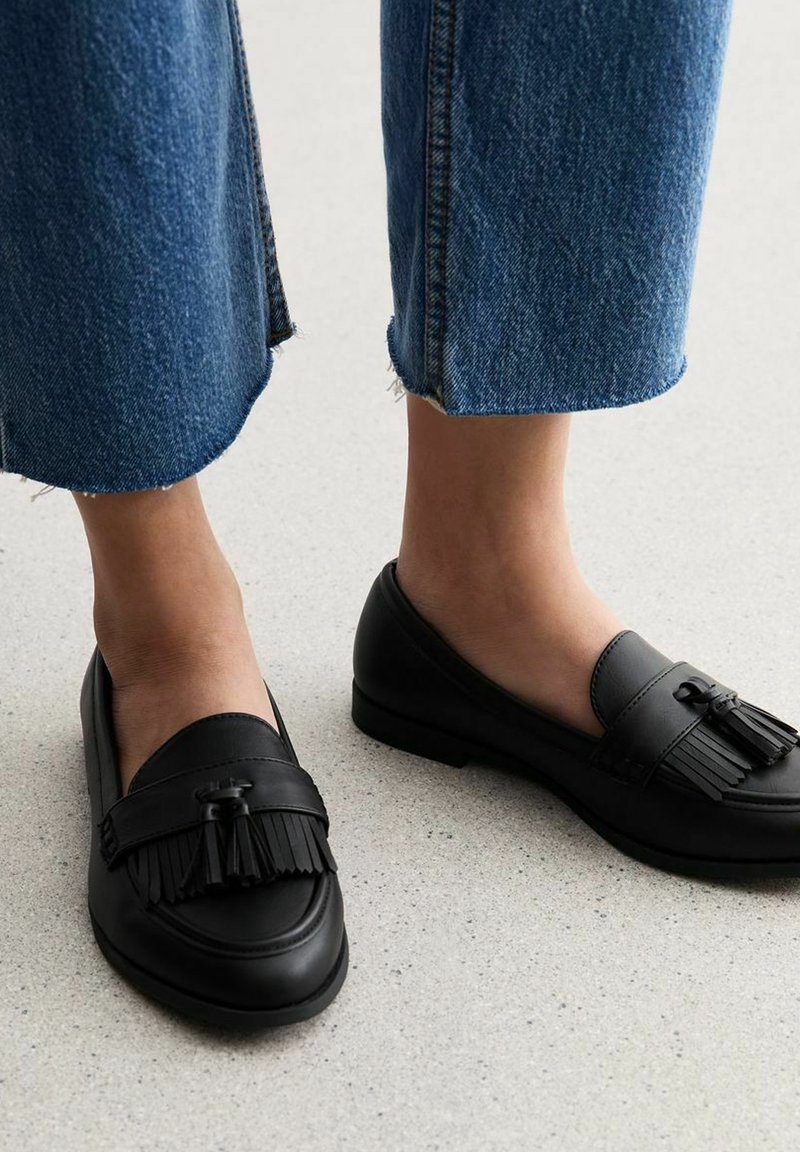 Mocassins en cuir noir à semelle plate, ornés d'un pompon décoratif et d'une finition lisse. Portés avec un pantalon en jean effiloché.