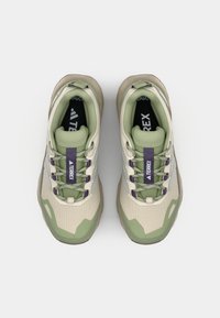 Beige og grønne joggesko med en teksturert stoffoverdel, lilla snøring og en polstret såle. Designet for utendørs bruk med en støttende struktur.