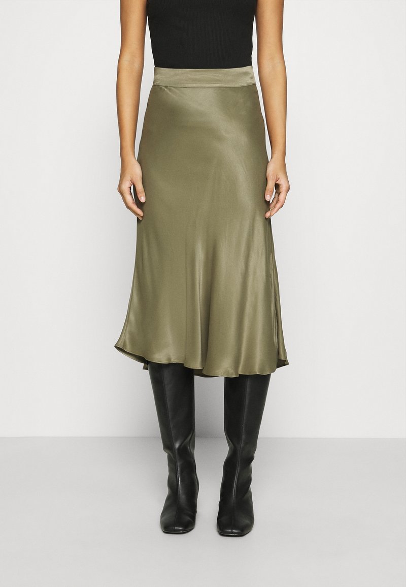 Femme portant une jupe midi en satin vert olive, un haut noir sans manches et des bottes en cuir noires montantes, debout devant un fond blanc uni.