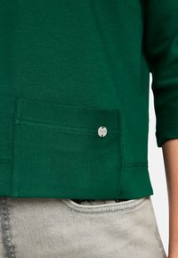 Grünes Baumwolloberteil mit Rundhalsausschnitt und drei Viertelärmeln, ausgestattet mit einer Fronttasche und einem silbernen knopfdetail.