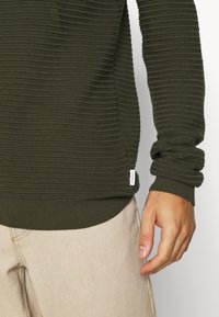 Mørkegrønn ribbestrikket genser med teksturert stoff, tettsittende ermer, og en liten merkelapp nederst på kanten, parret med lyse beige bukser.